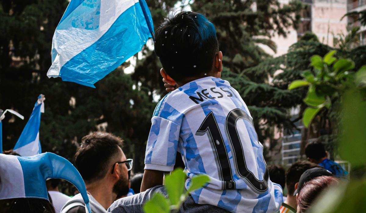 A child on the shoulders of a parent wears a light blue and white stripped football top, waves the Argentinian flag