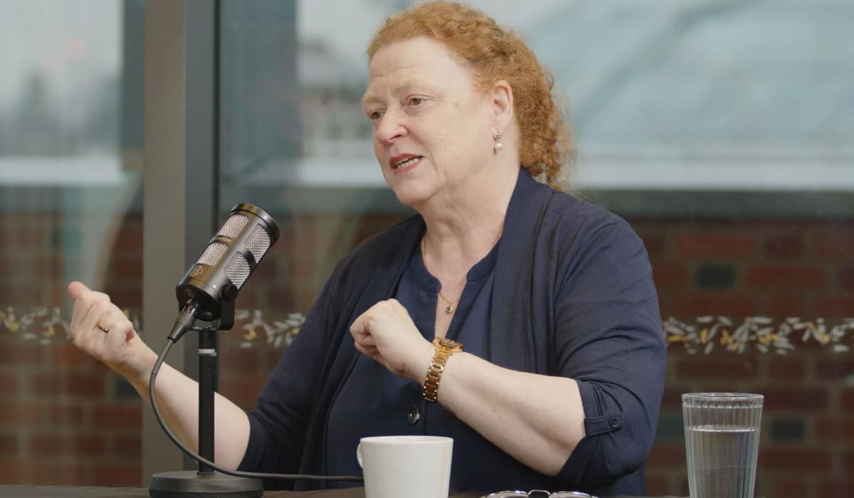 A woman guest on a podcast sits at a table with a mic in front, gesturing to the side with both hands.