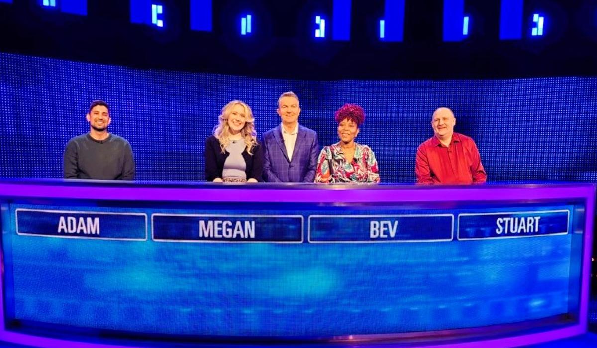 Four quiz show contestants stand behind a table with the show host.