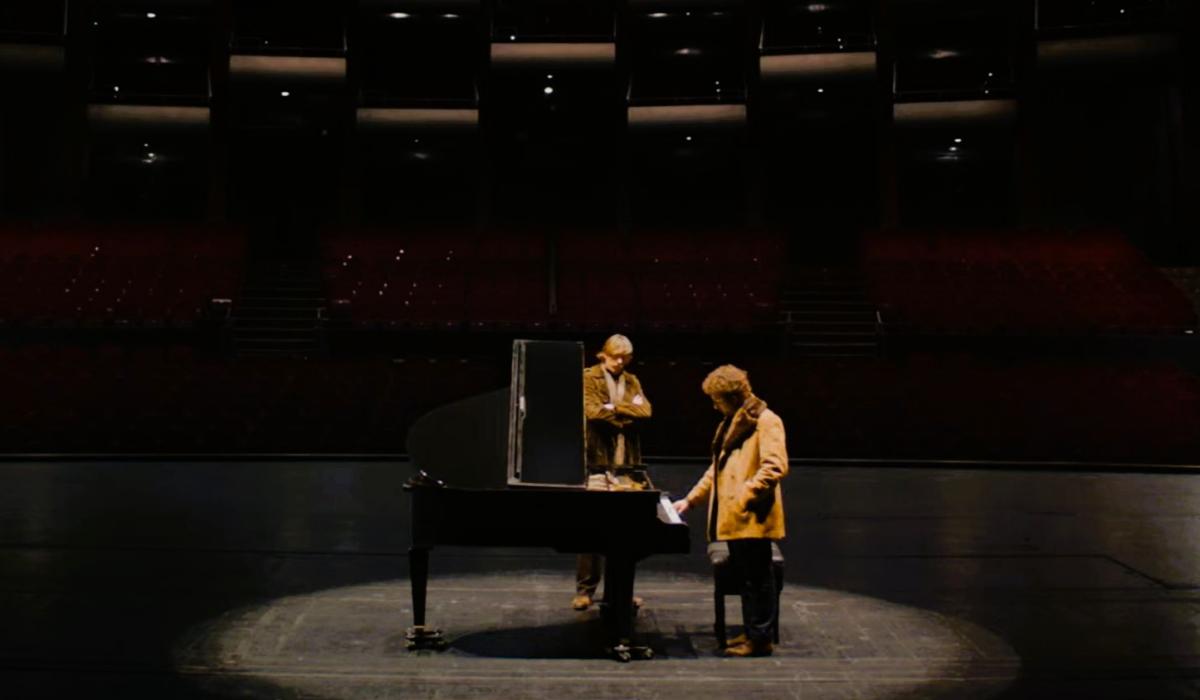 Two people sit and stand next to a grand piano on a stage.