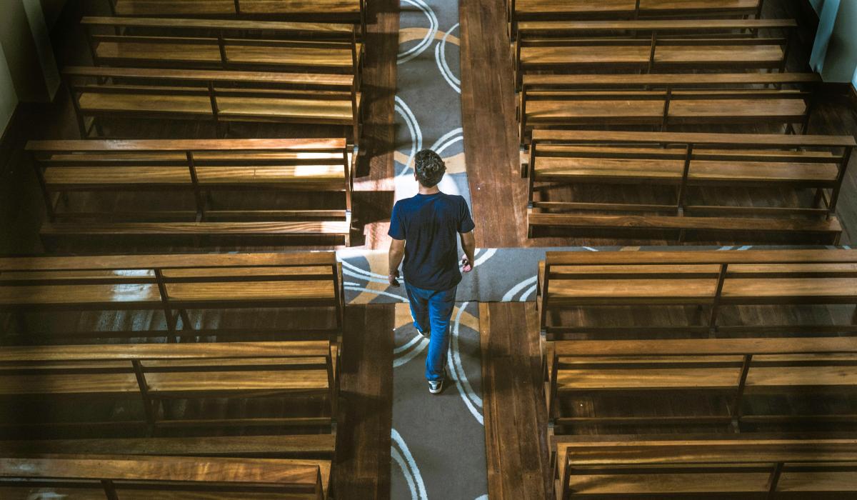 A casually dressed man strides down the aisle of a church between the pews.