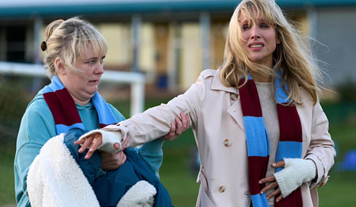 On the sidelines of a pitch a well-dressed mum hands a coat to a sceptical looking mum beside her.