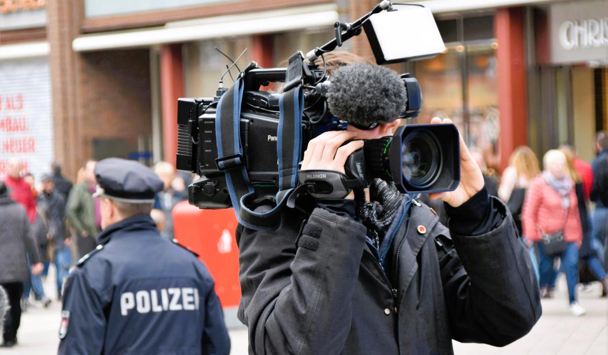 A news cameraman holding a camera, stands back to back to a police officer.