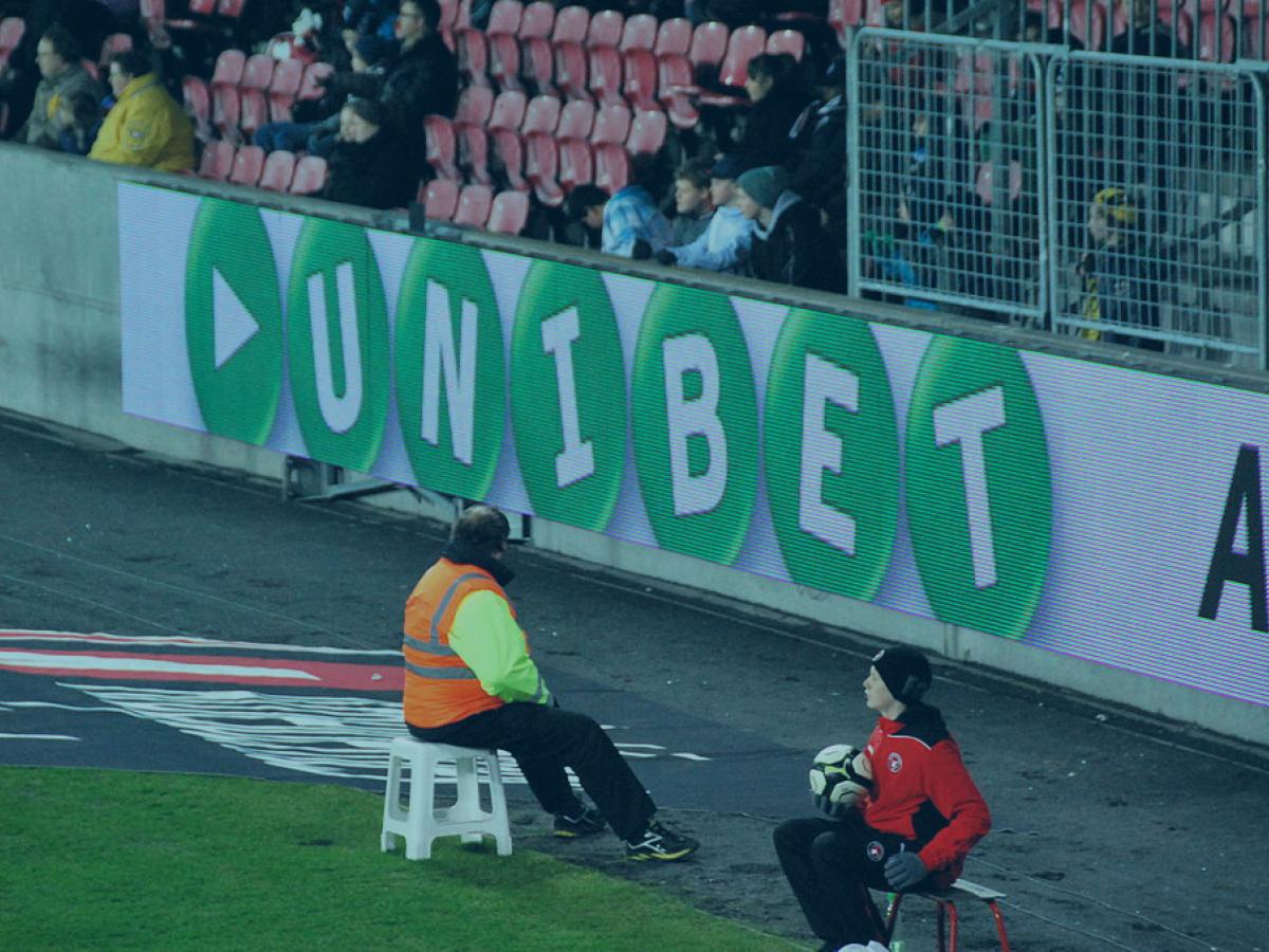 The edge of a football pitch showing an advertising hoarding with a betting brand name on it.