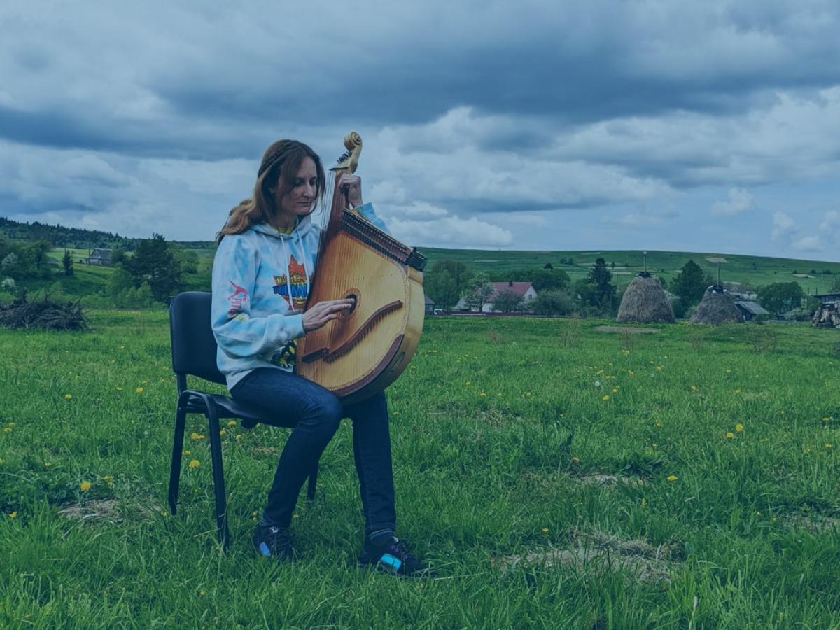 A woman sits on a chair in a field holding a large stringed musical instrument.