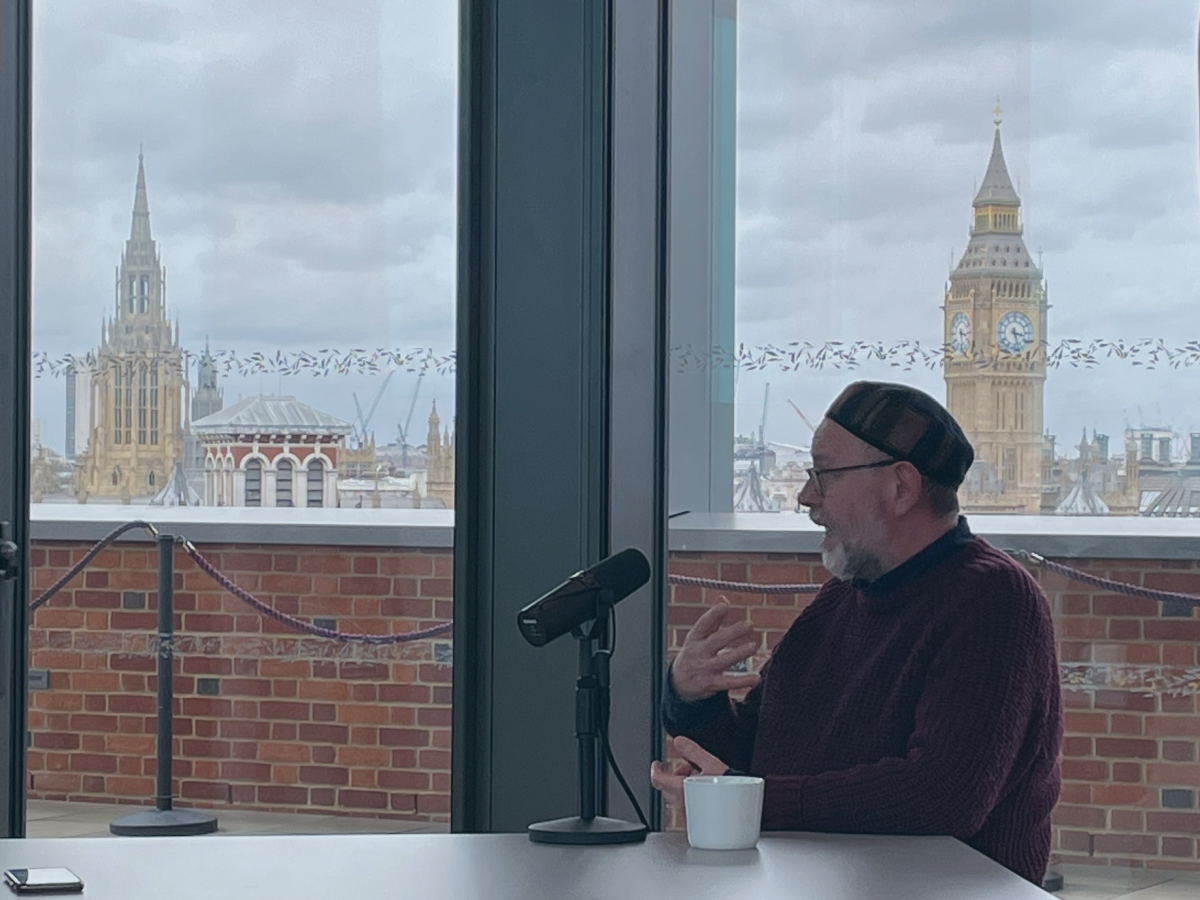 A man sits and speaks into a microphone beside him on a table, while gesturing with his hand. Behind, Big Ben is on the skyline.