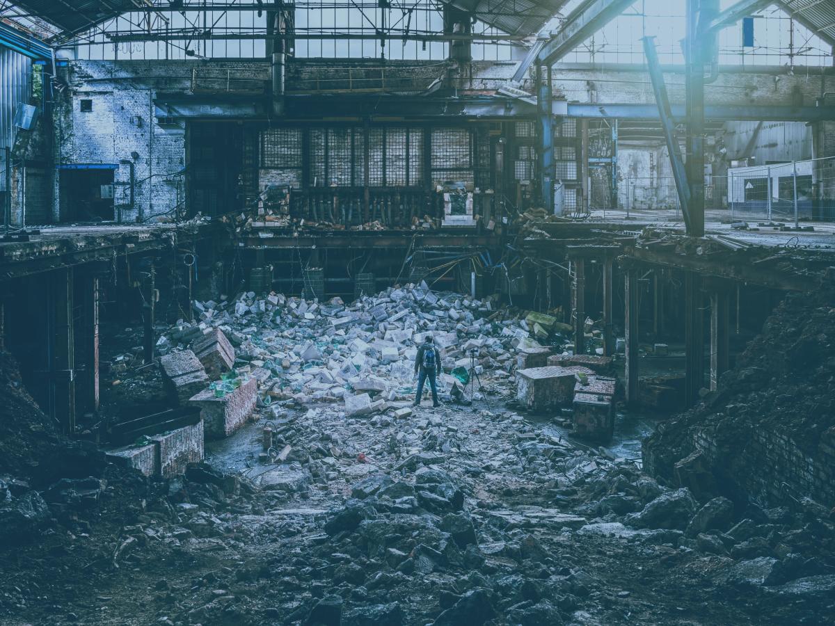 A photographer, standing next to a tripod, atop a pile of rubble is a destroyed factory.