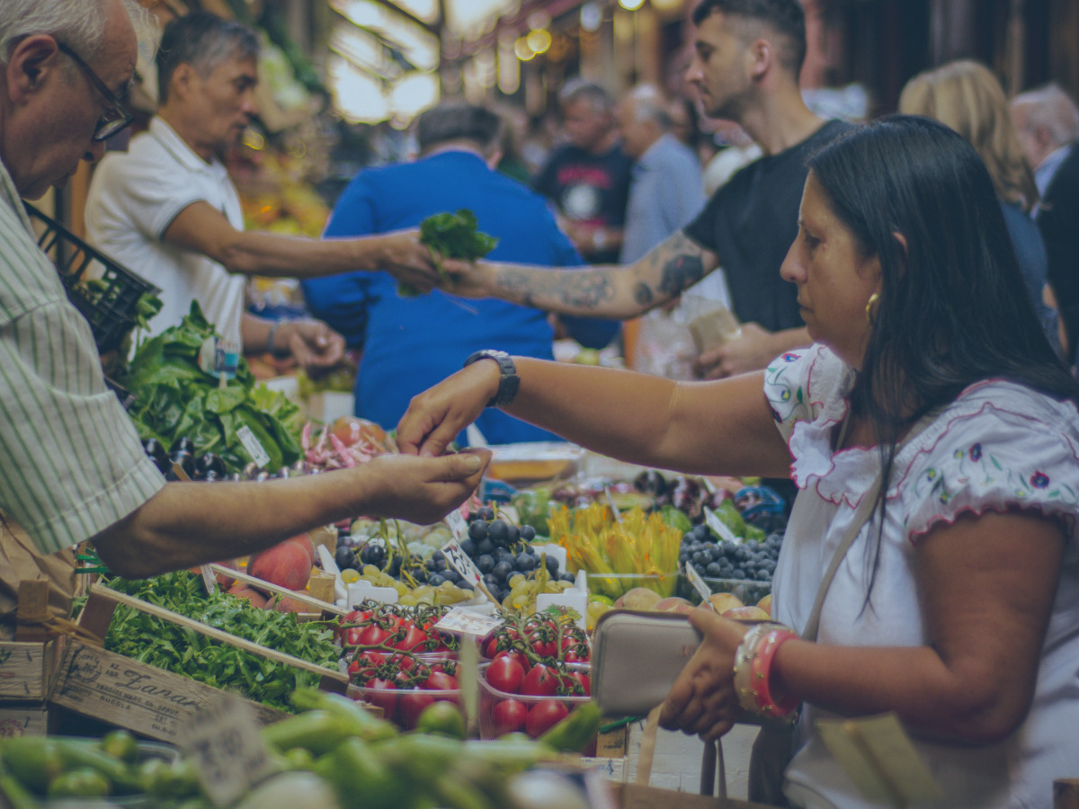 Marketer stall traders and customers reach out arms and hands to exchange fruit and money above piles of produce.