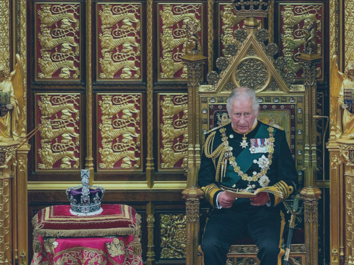 A uniformed Prince Charles sits on a throne reading a speech, beside a crown resting on a cushion