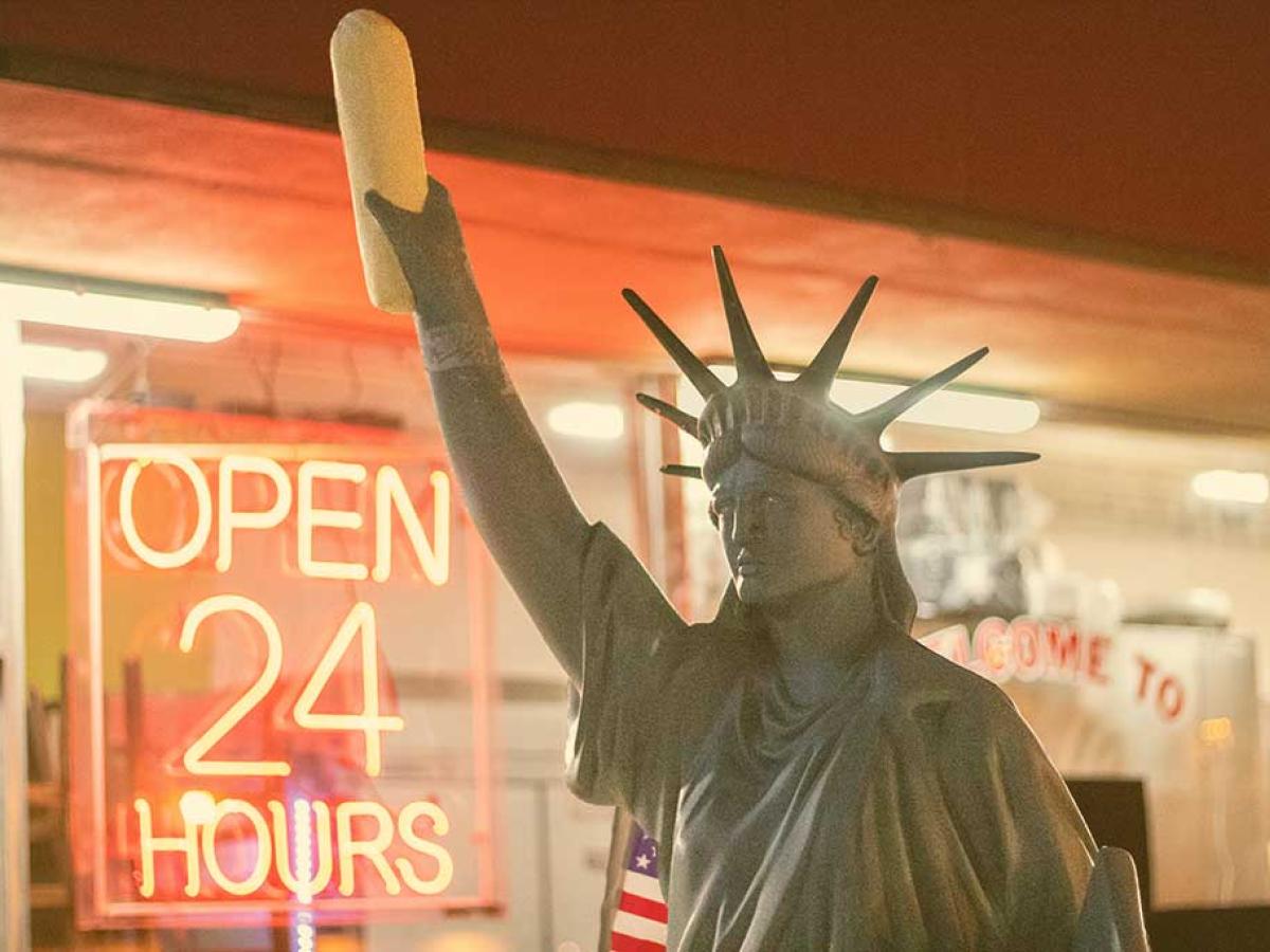 A copy of the Statue of Liberty, holding a stick of bread, stands outside a shop window displaying an 'Open 24 Hours' signs.