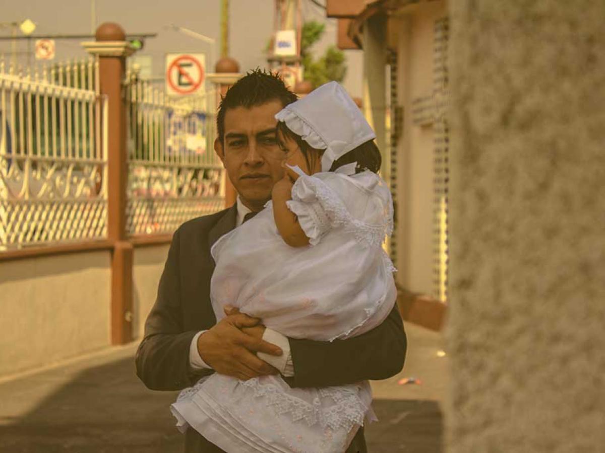 A father wearing a suit carries his child who is dressed for a christening in white.