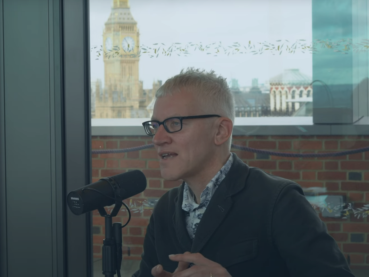 A man sits at a table speaking into a microphone but looks into the distance. Behind him, through the window and beyond a wall is Big Ben