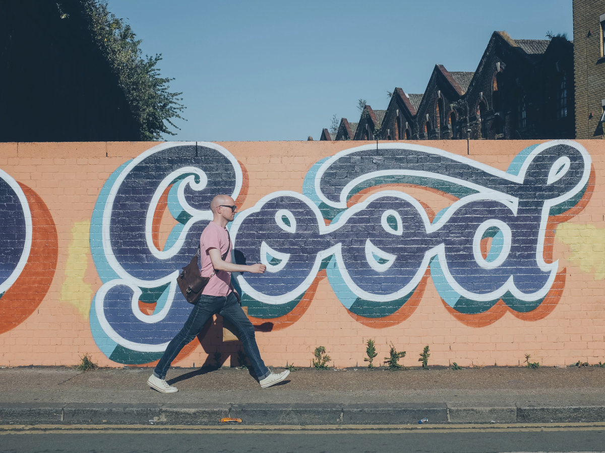 A man walks along a street past a orange wall with a huge 'Good' written in cursive script on it.