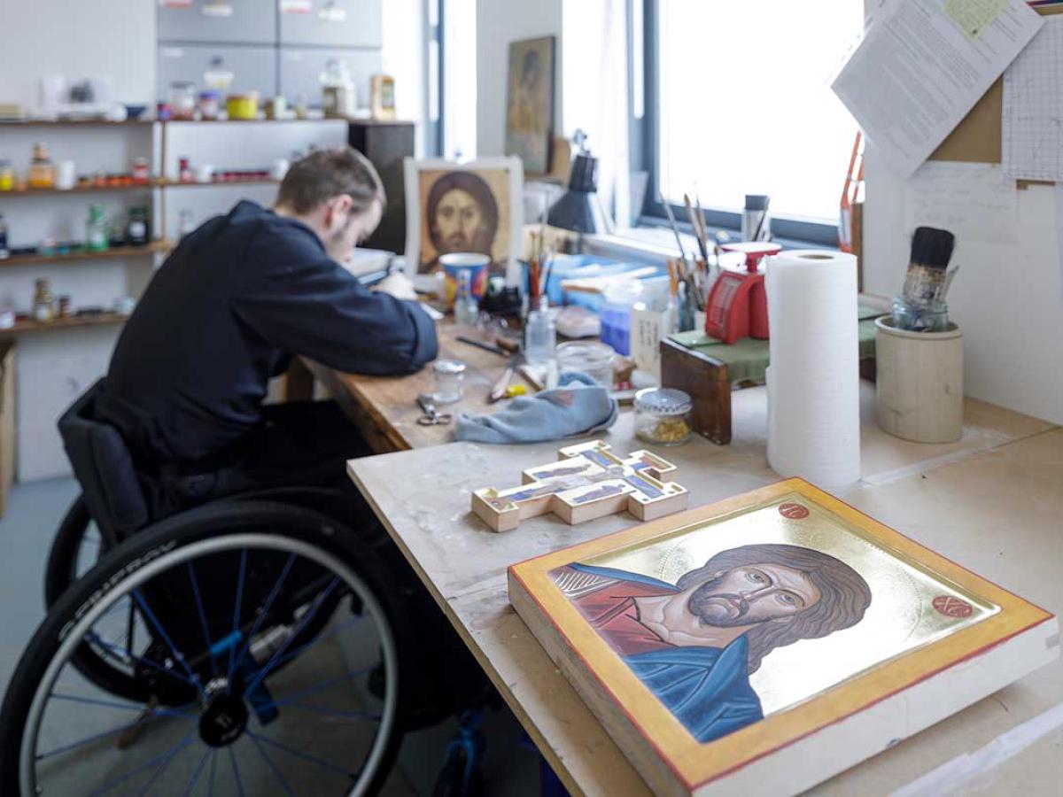 A monk in a wheekchair works on an icon in an art studio. In the foreground is a completed icon. 