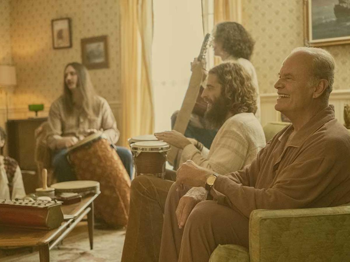 A group sit in a lounge playing musical instruments while the man closest to the camera laughs.