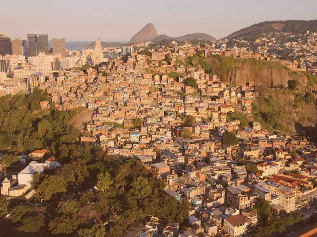 an aerial view of a shantytown on a steep hill side in Rio, Sugerloaf Mountain is visible in the distance 