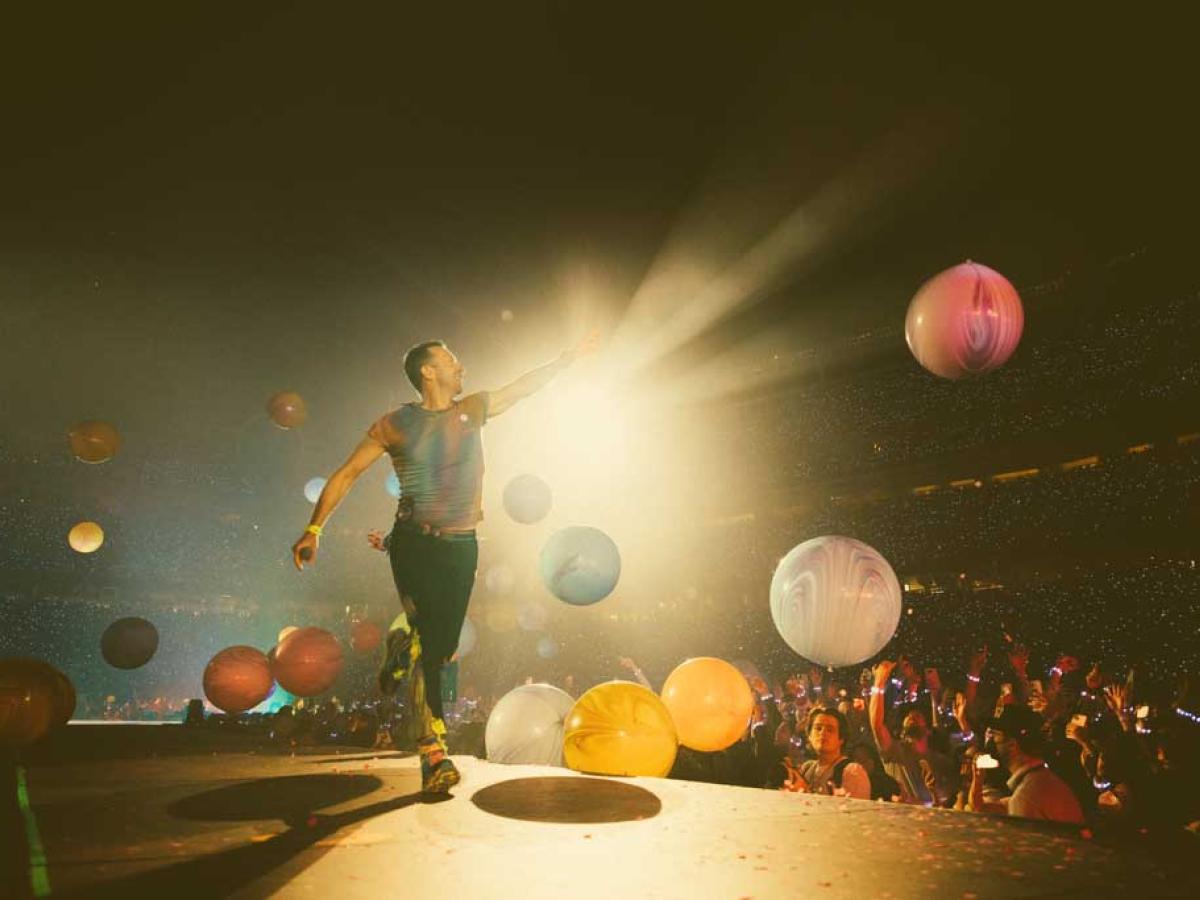 A singer struts a stage pointing to the spotlight as coloured orbs float down.