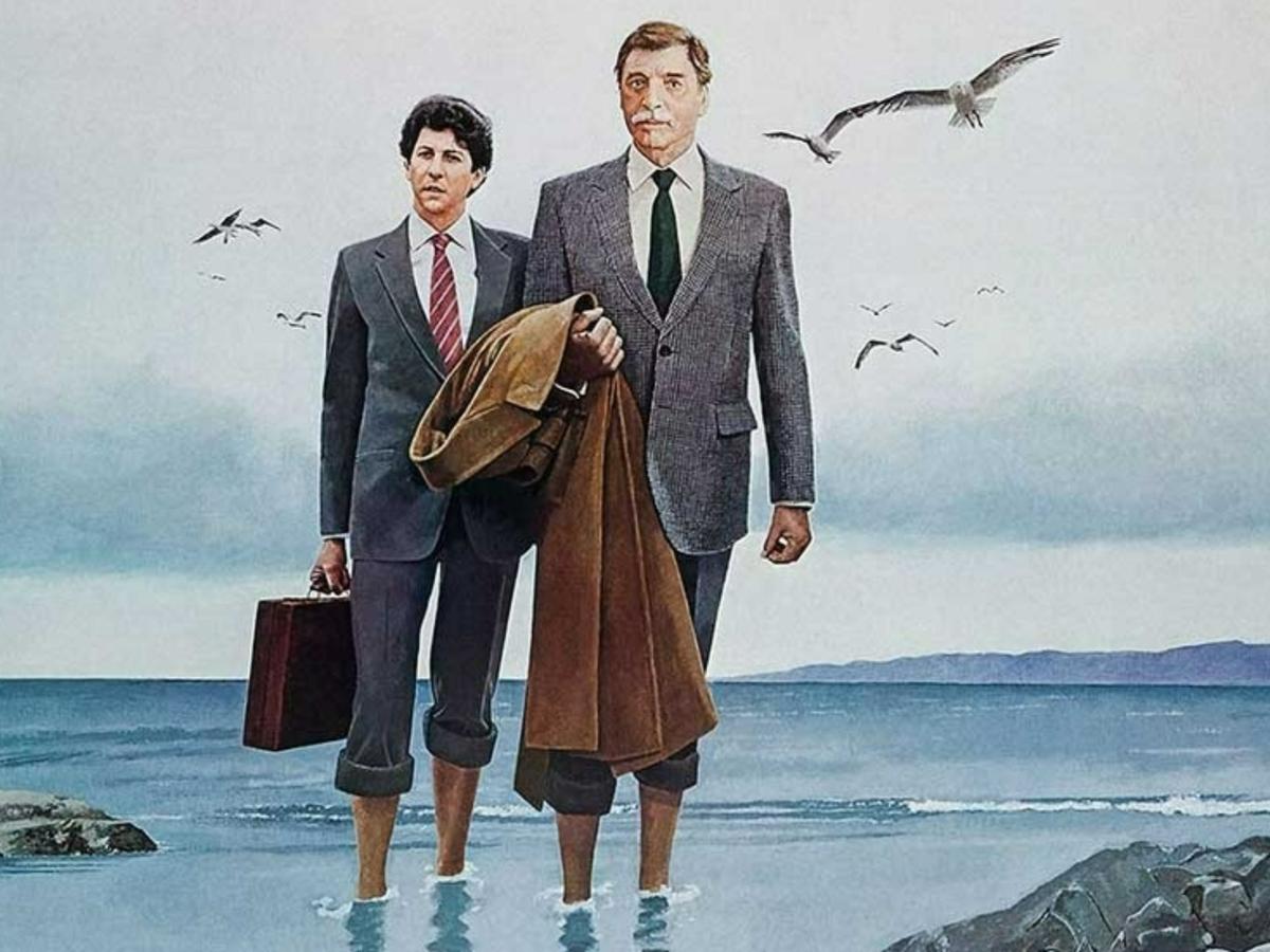 Two business men in suits hold coats and briefcases, stand in the sea with their trousers rolled-up above their ankles