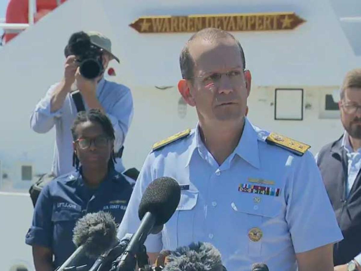 A Coast Guard officer gives a press conference while looking grim-faced. Others look on.