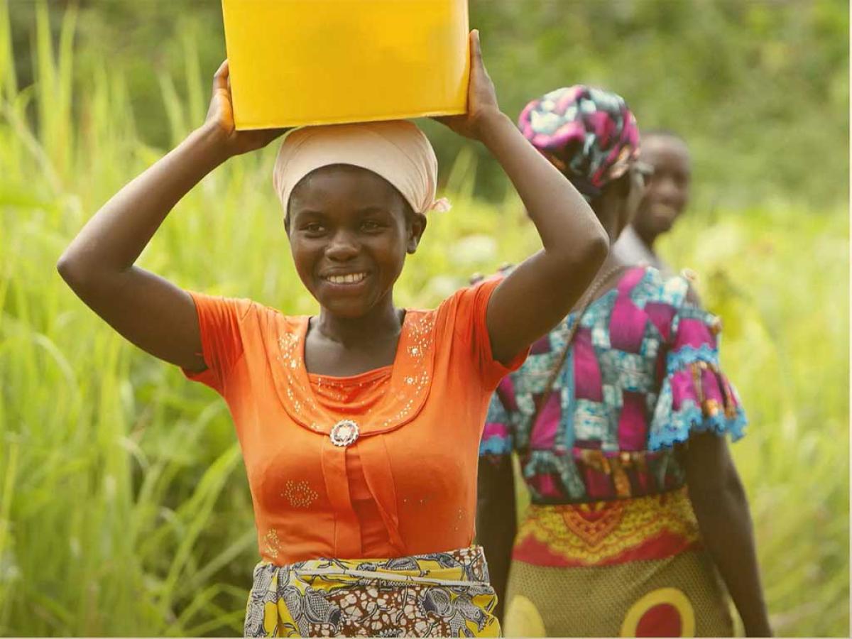 A woman carries a plastic bucket on her head and held by raised arms and hands.