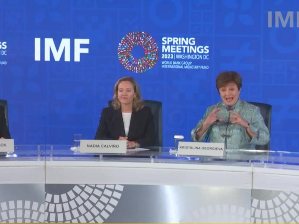 Two women sit behind a press conference desk against a backdrop, one listens as the other speaks and  gestures.