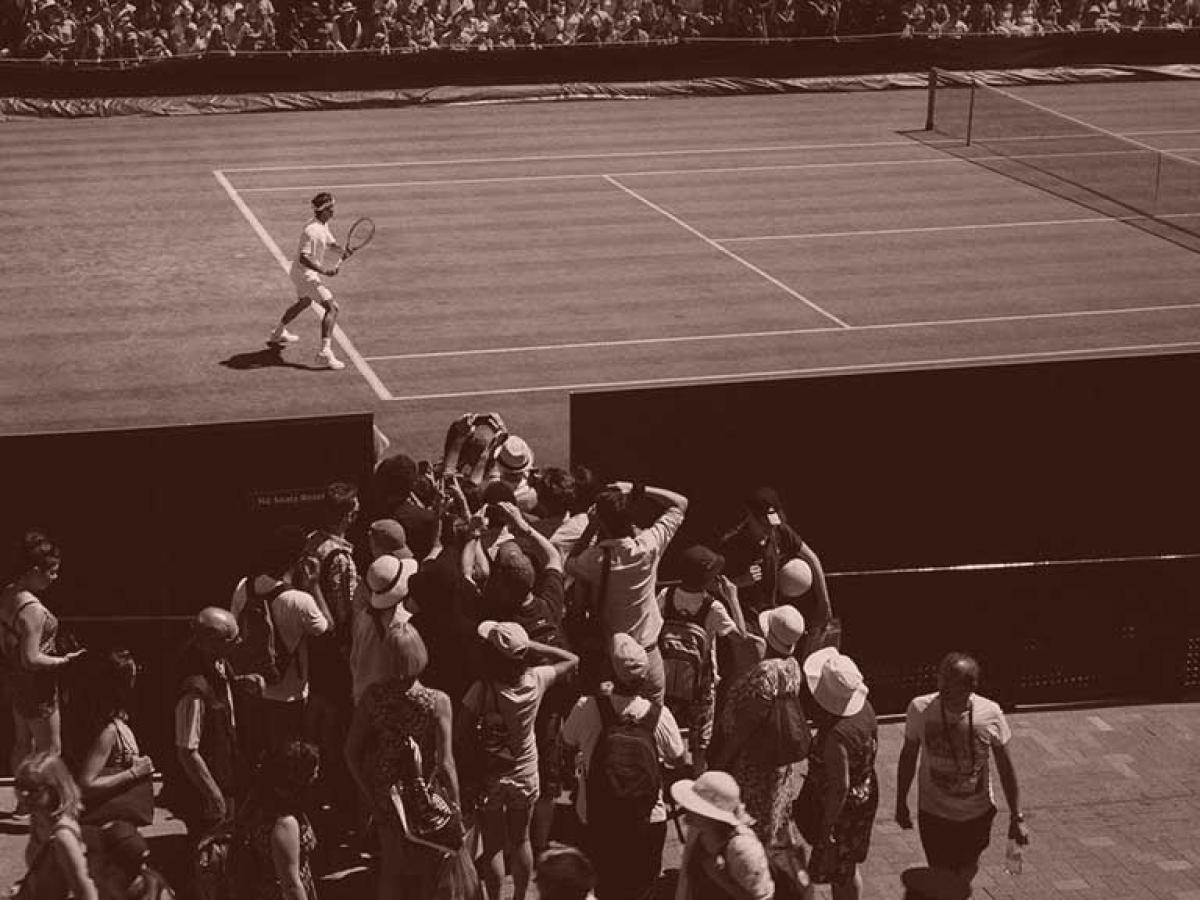 A tennis player stands ready to return a shot, while a phalanx of photographers crowd round a court-side opening to take a picture of him.