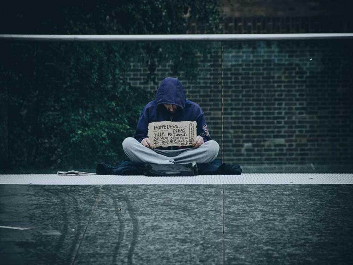 A beggar sits cross legged against a glass railing holding a sign.