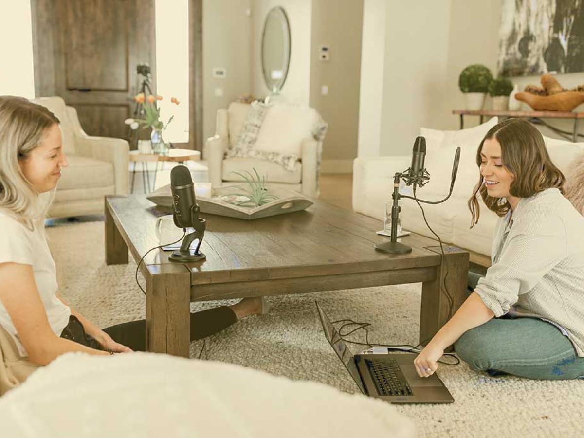 Two people sit cross-legged at a low table on which two microphones stand. One press a key on a laptop on the floor.