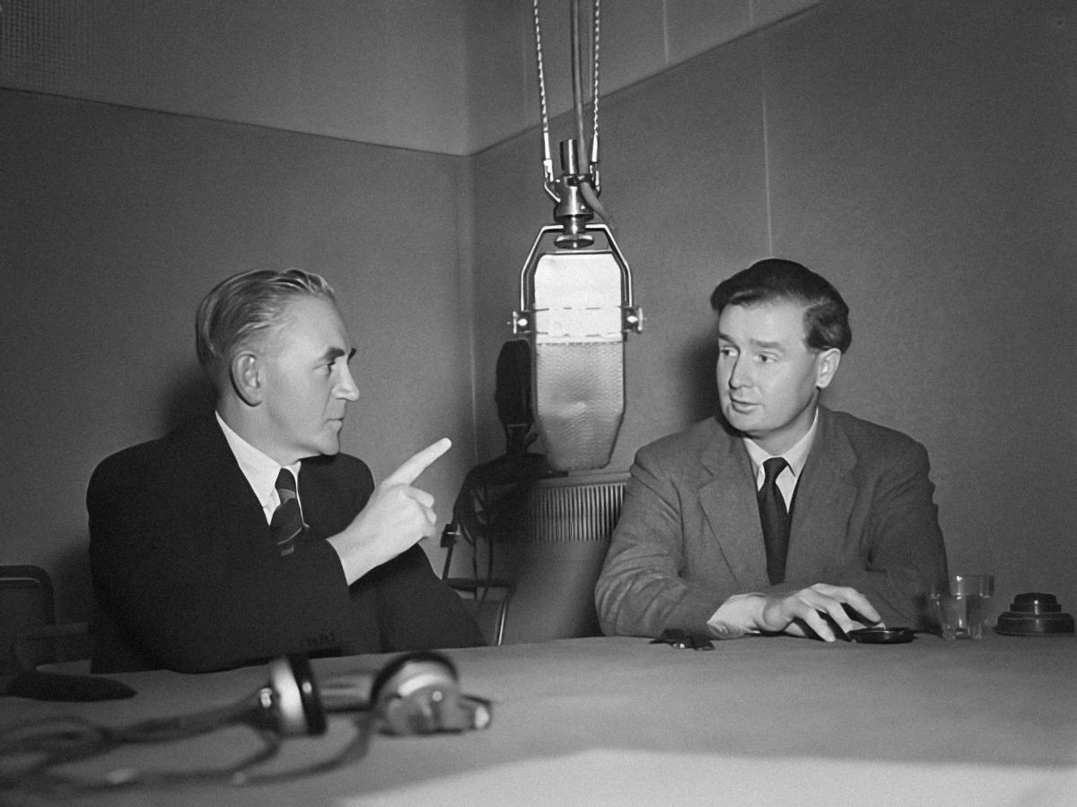 Two 1950's men un suits sit at a table dominated by a large hanging microphone. One points a raise hand and finger into the air. The other listens.