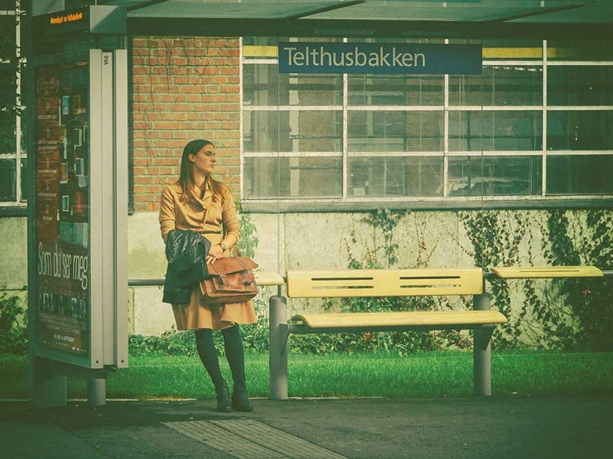 A woman leans against the glass of a bus shelter while waiting, she clasps a bag.