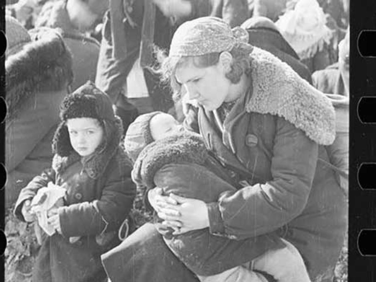 A mother cradles a child while another stands close by. They wear winter clothes of the 1940s and are amidst others waiting.