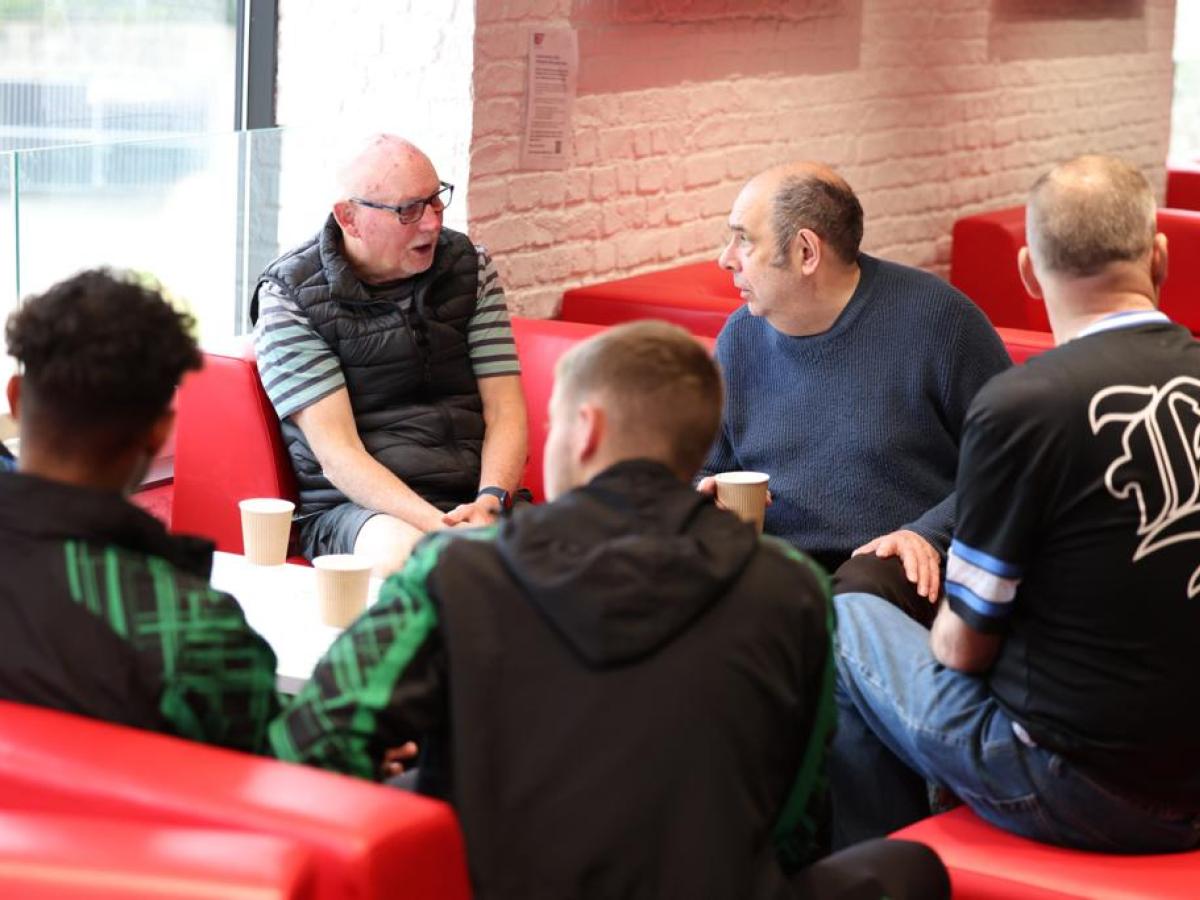 A group of old and young men sit on red sofas, listening to one of themselves.