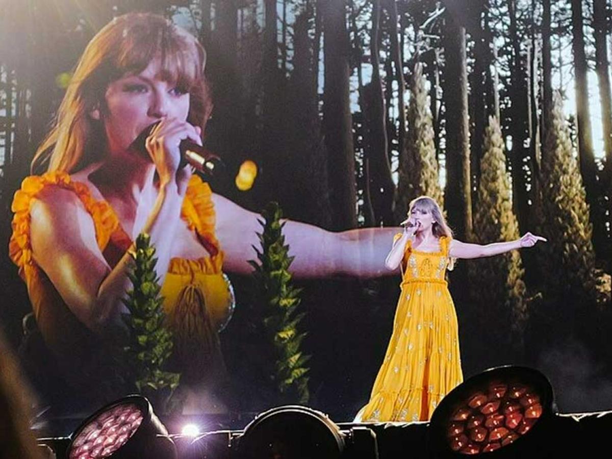 Taylor Swift wears a long yellow dress and signs with an outstretched arm against a backdrop of woods and a close up her singing beside it.