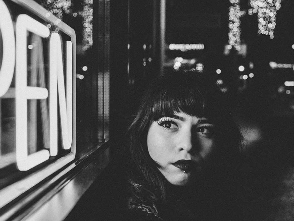 A black and white close up of a women in a street at night, turning to look around at a neion 'open' sign.