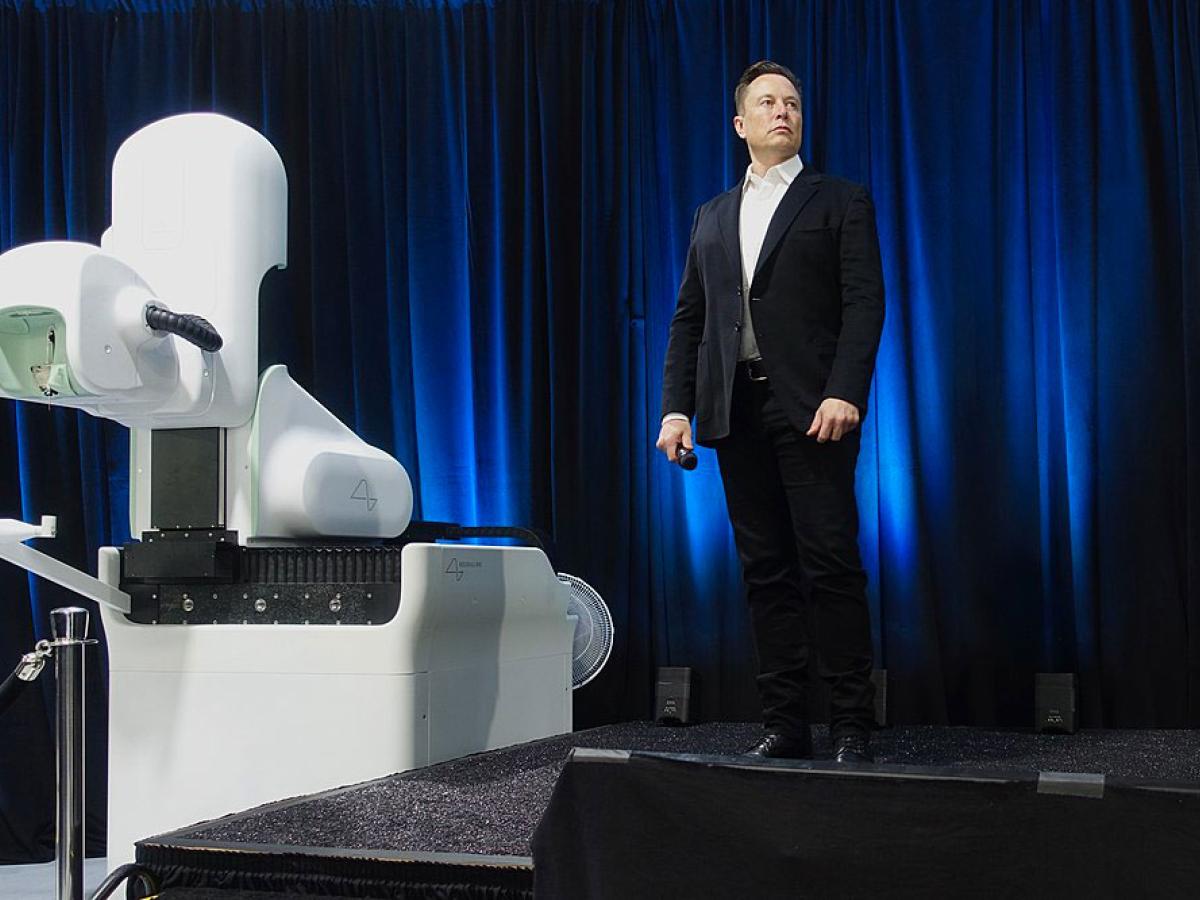 Elon Musk, wearing a dark suit, stands on a stage to a white robotic looking surgical robot.