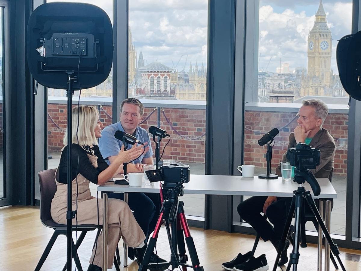 Around a table with microphones, three people record a podcast, one leans in talking and gesturing with a hand while the others listen.