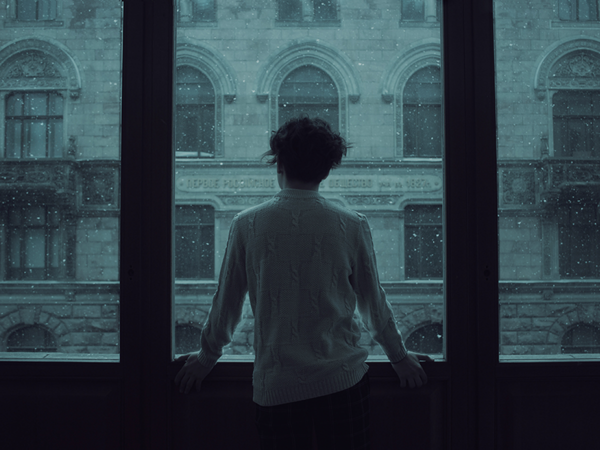 A person stand looking out a wide set of windows covered in rain.