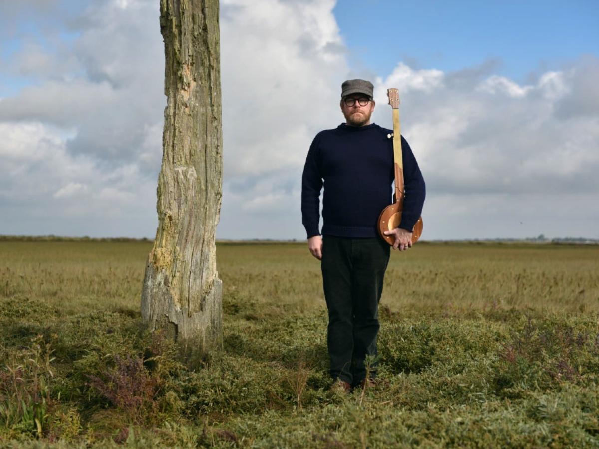 Standing on a salt marsh beside a wooden pillar, a man holds a banjo upright like a rifle.