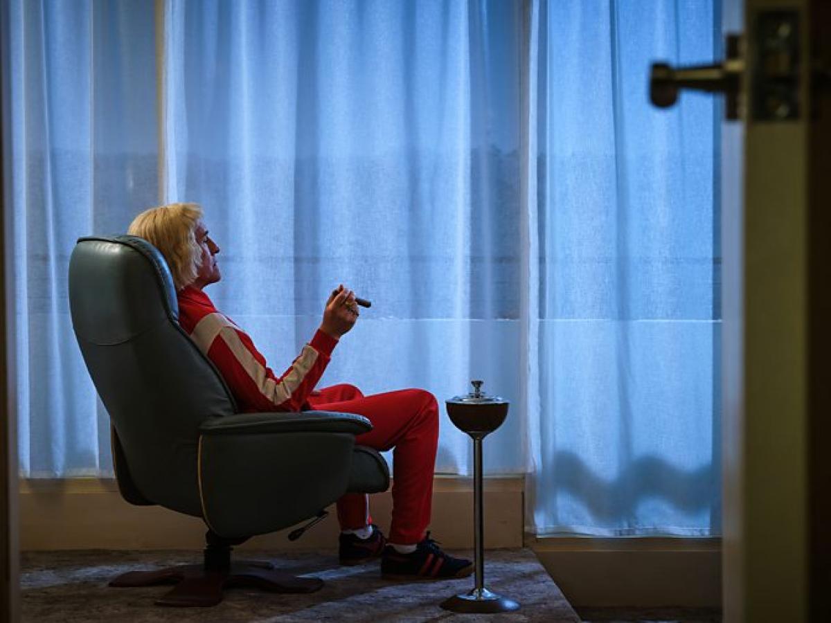 A man in a tracksuit sits in an arm chair smoking a cigar while looking towards a curtained window.