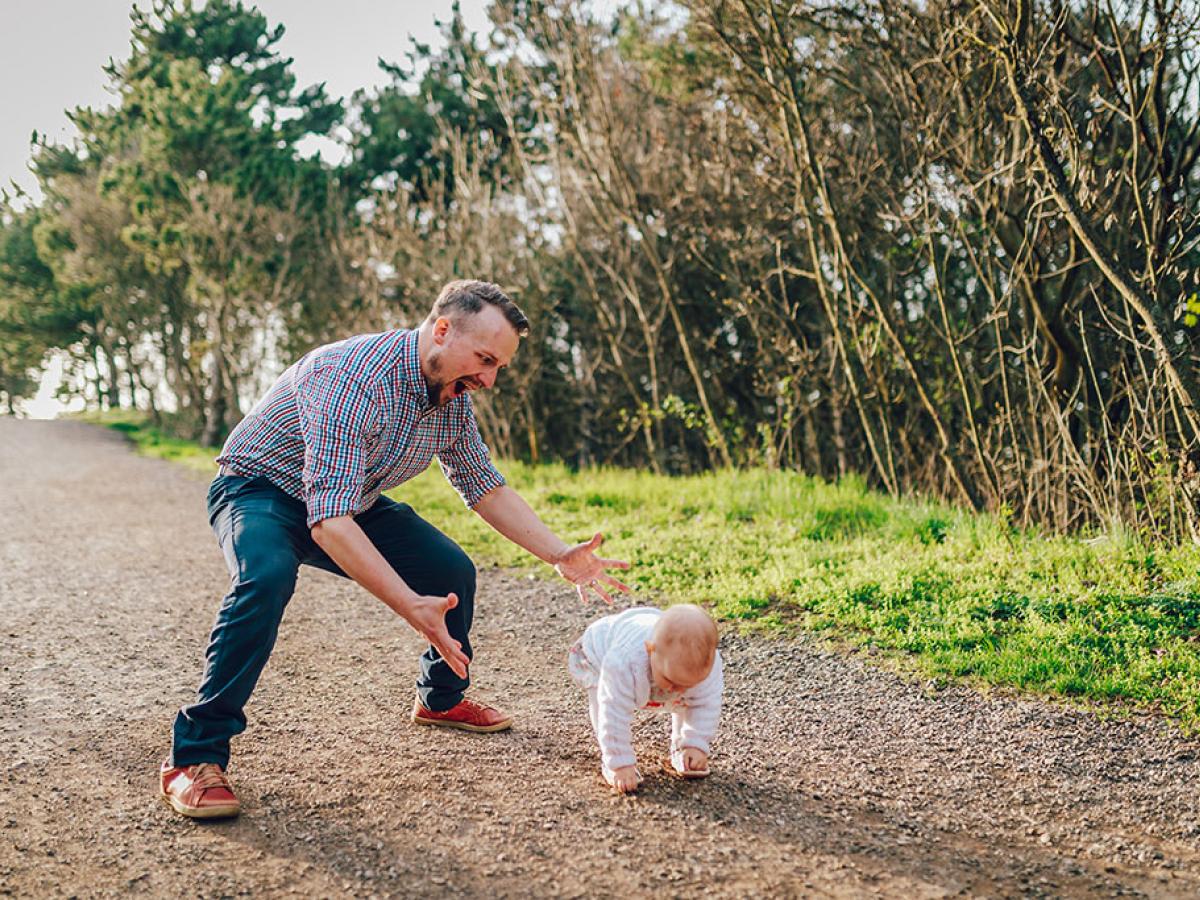 A dad hovers with open arms ready to catch a baby taking first steps