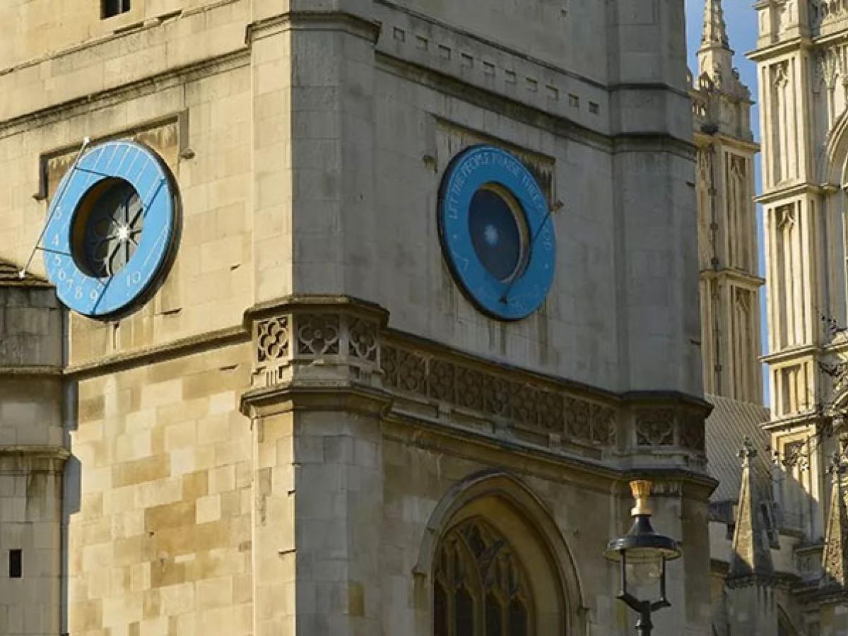 A close up of a blue sun dial on a church clock tower, with an abbey's windows behind it.