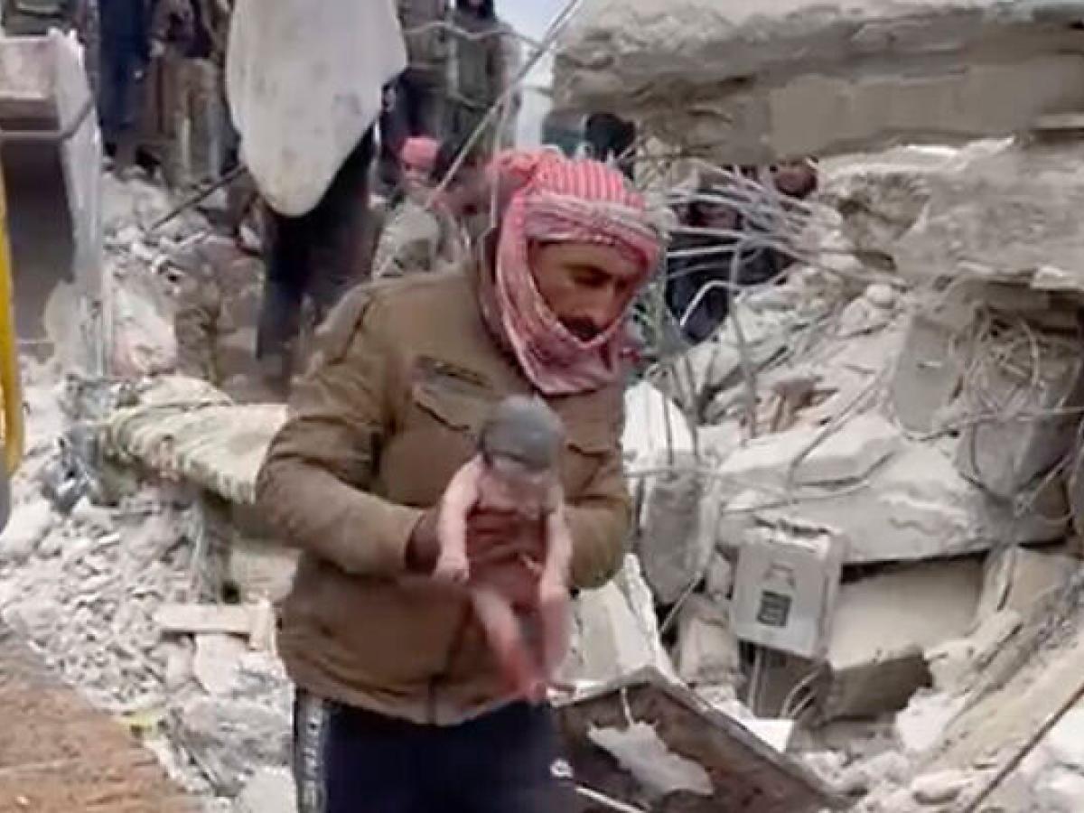 In front of a collapsed building, a rescuer carries a new born baby by the arms.