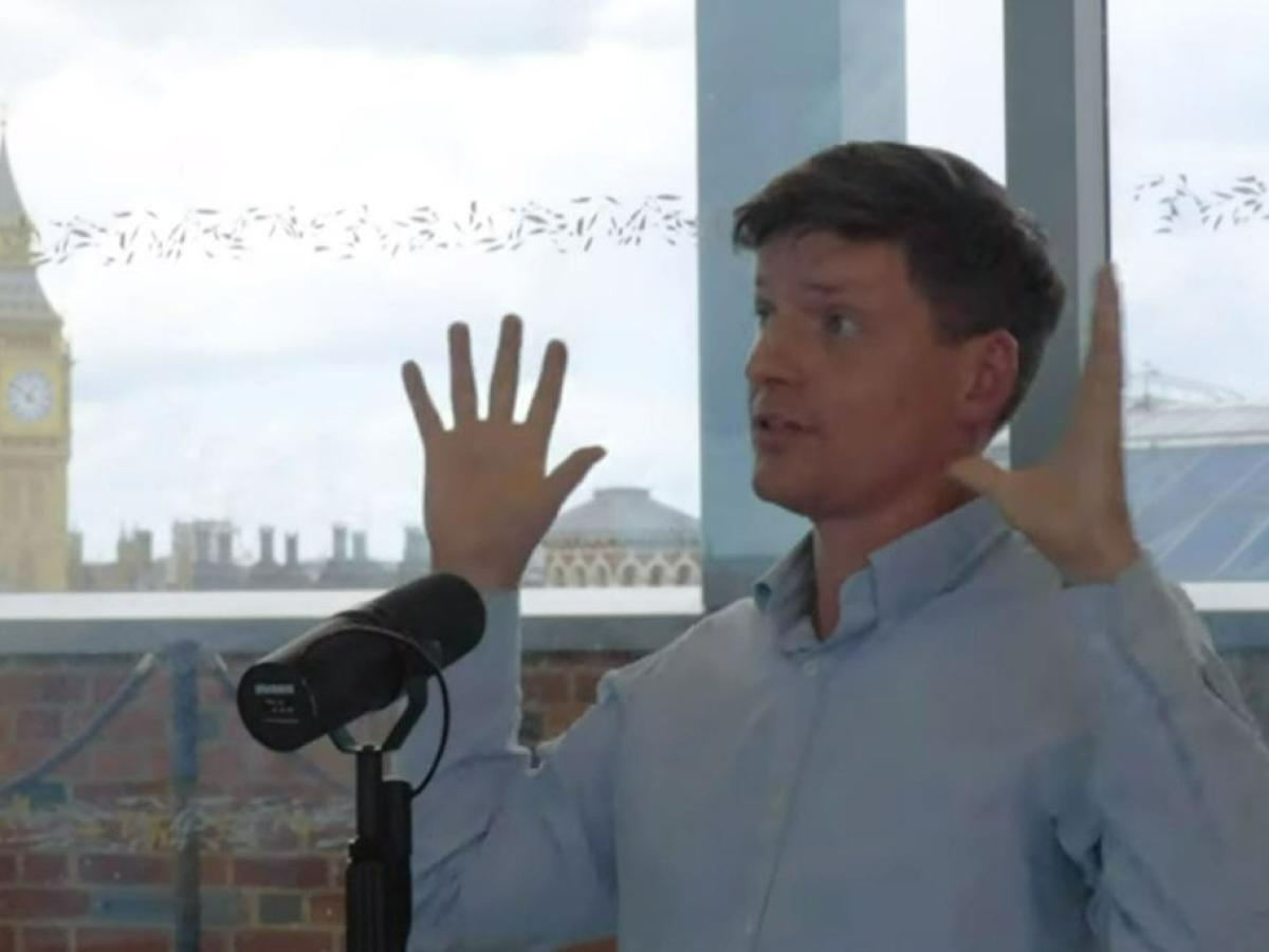 A man sits at a table recording a podcast and holds his hands up for emphasis.