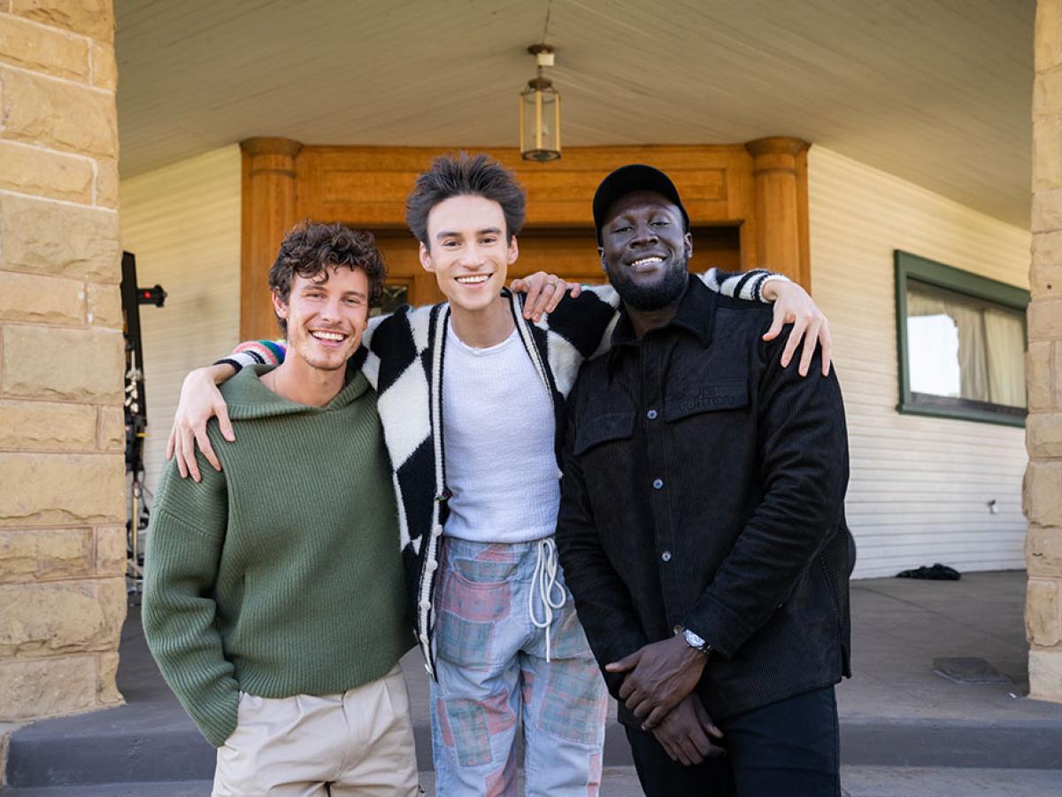 Three happy men stand with the one in the middle draping his arms around his friends.