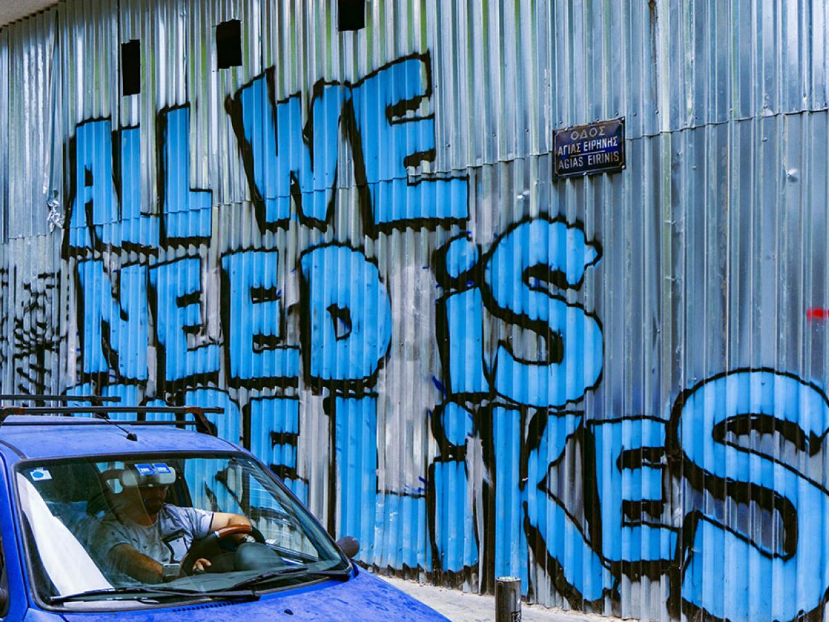 A corrugated sheet iron wall graffited in large blue letters that say 'All we need is more likes.'