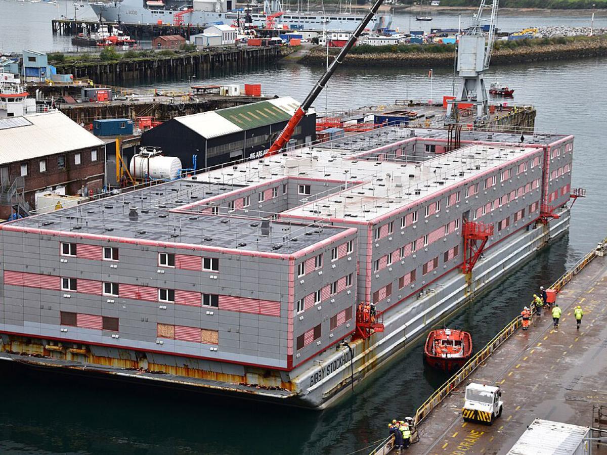 A grey multi-story accommodation barge floats beside a dock.