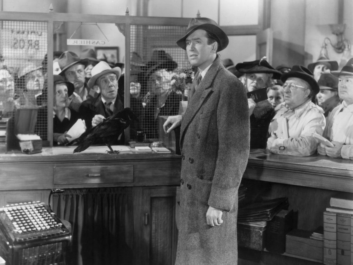 A man stands one side of a bank counter while others, on the other side, look hopefully at him.