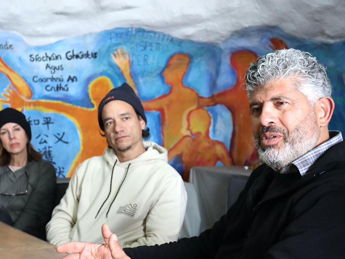 Two people down a table turn and listen to someone closer talk, against a wall mural.