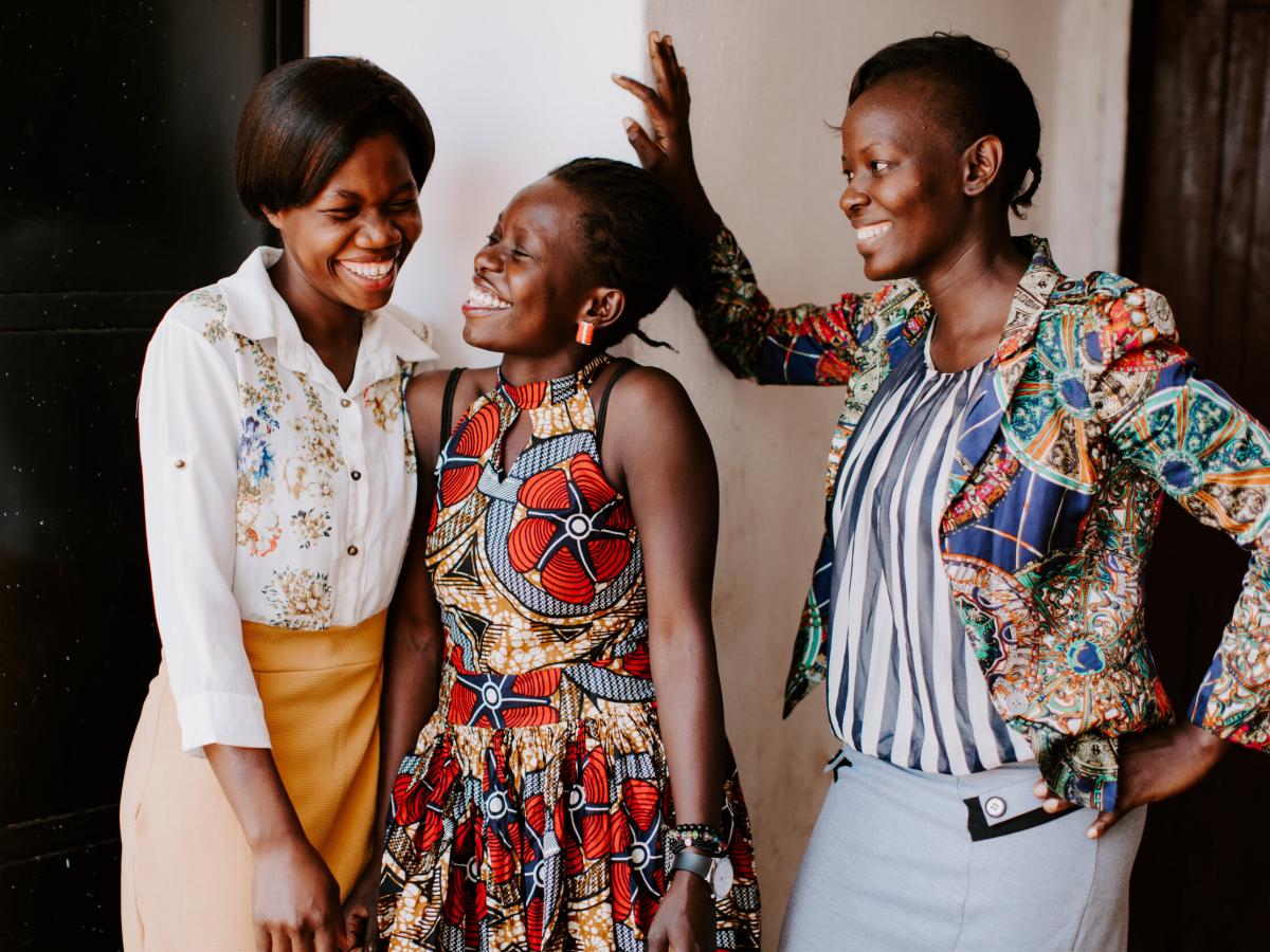 Three women stand, two lean into each other sharing a joke, while the other laughs too.