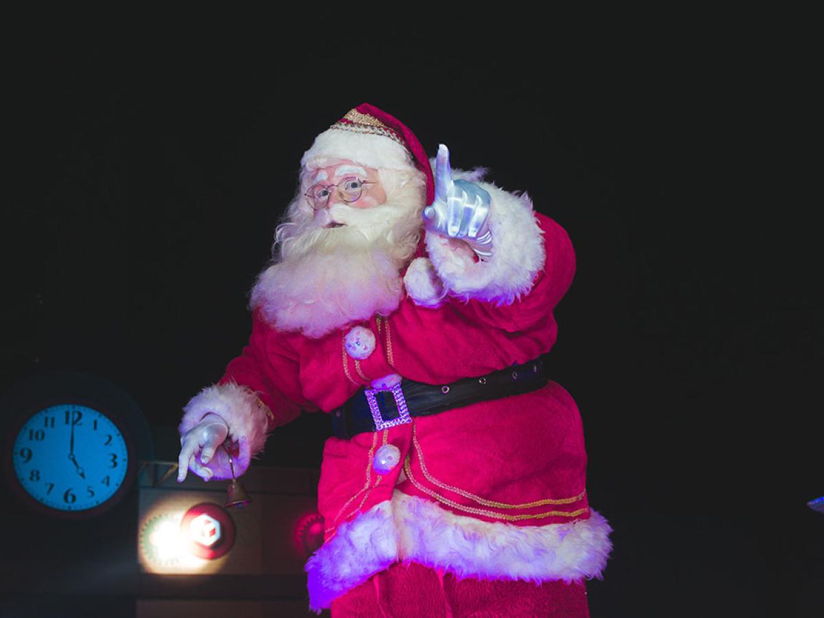 A Santa tiptoes in a darkened room carrying a lamp and holding up a finger in a gesture of silencing